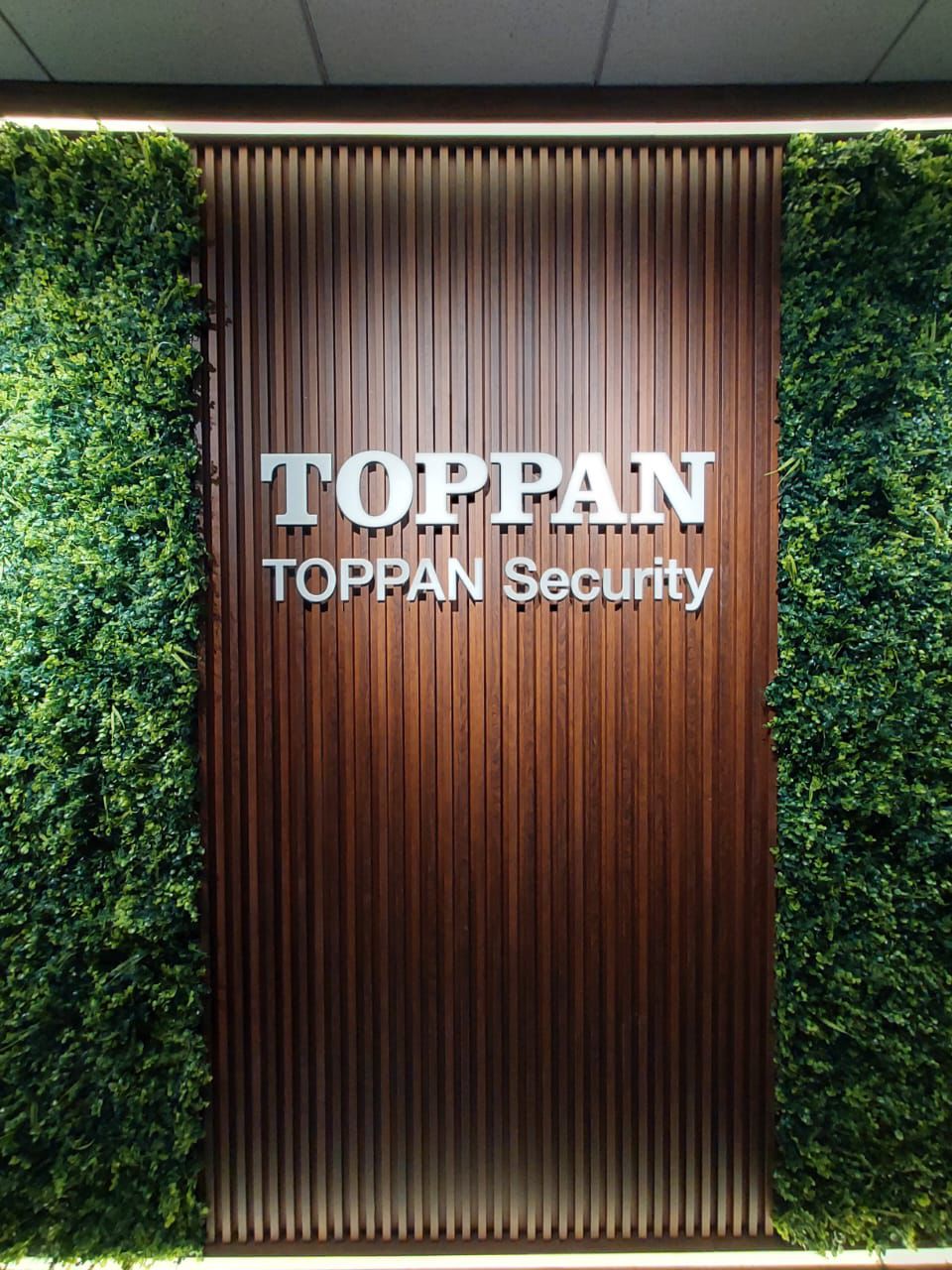 3D stainless steel corporate signage for TOPPAN Security mounted on wood panel and green wall Dubai